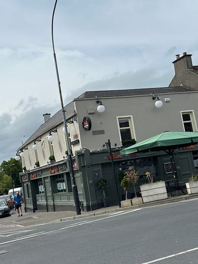 The image depicts the exterior of "Flame Steak House" located in the city of Carlow, Ireland. The building is a two-story structure with a light grey facade on the upper level and a dark green lower level that houses the restaurant. Several round, white outdoor lights are mounted on the upper facade. A green umbrella shades an outdoor seating area in front of the restaurant.
On the sidewalk in front of the restaurant, a man with a beard and blue t-shirt is jogging towards the right side of the frame. He is wearing dark shorts and athletic shoes. Several cars are parked or driving on the street in the background. The sky is overcast with grey clouds. A lamppost is prominently positioned in the foreground, leaning slightly to the left. A chimney with a brown cap is visible on the roof of the building. The street in front of the building appears to be a public road with white lane markings. The overall impression is a daytime street scene in an urban environment.