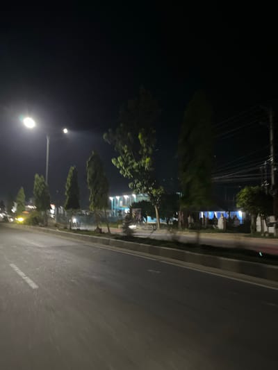 The image shows a street scene at night in Tanjungkarang, Indonesia. A road occupies the right side of the frame, with a white line marking the lane. To the left of the road, there is a sidewalk lined with trees and streetlights. In the background, buildings with illuminated windows are visible, suggesting an urban or semi-urban environment. The sky is dark, indicating nighttime. The overall impression is one of a quiet street at night, possibly in a residential or commercial area. There are no people or vehicles clearly visible in the image, and no text is legible.