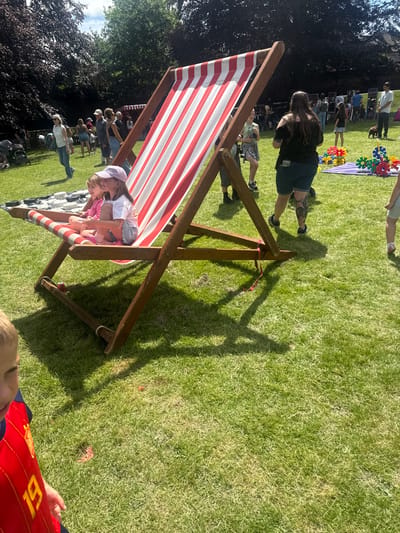 The image depicts a sunny outdoor event taking place in the city of Leominster, United Kingdom. In the foreground, a large red and white striped deck chair is prominently featured, with two young girls sitting in it. The girl on the left, wearing a pink top and a pink cap, is smiling. The girl on the right, in a white top and shorts, is looking to her right. The deck chair is situated on a well-maintained green lawn.

In the mid-ground, a variety of people are visible, engaged in different activities. Some are standing and conversing, while others appear to be browsing. Behind them, a red and white striped stall or tent is visible, suggesting a market or fair-like atmosphere. To the right of the stall, a collection of colorful plastic building blocks are arranged on a purple mat. Further back, more people are spread across the lawn, and in the distance, trees line the perimeter of the area.

The overall scene suggests a lively community event, possibly a summer fair or festival, with families and individuals enjoying the pleasant weather and various attractions.