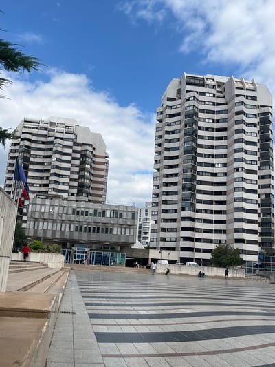 The image shows a view of the Place de l'Échutier in Créteil, France, with two tall, modern buildings dominating the skyline. The building on the right is a multi-story residential tower with a distinctive facade featuring numerous balconies and windows. The building on the left, though shorter, also exhibits a similar architectural style with multiple levels and windows.
In the foreground, a wide plaza with a distinctive black and white striped pavement pattern extends towards the buildings. To the left of the plaza, there are concrete steps and a ramp leading up to the entrance of one of the buildings. Two flags, appearing to be the French flag and another national flag, are displayed on flagpoles near the entrance.
Several people are visible in the scene. On the steps to the left, a person in a red shirt is ascending. In the plaza, individuals are seen walking, sitting, and interacting. Some are gathered near the entrance of the building, while others are further out in the plaza. A person on a scooter is visible in the middle ground. The sky above is blue with scattered white clouds. The overall environment suggests an urban public space with residential and possibly commercial buildings.