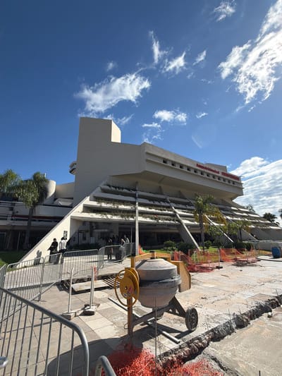 The image shows the exterior of a large, tiered concrete building, possibly an airport terminal, with red lettering spelling "Aeropuerto Internacional SILVIO PETIROSSI" visible on the upper level. The building has a distinctive modern architectural style with angled concrete supports and integrated greenery on the terraces. In the foreground, there is construction activity, indicated by orange safety netting, a concrete mixer, and excavated areas with visible rebar. Several people are standing near the entrance, some appearing to be waiting in line or in conversation. The sky is blue with scattered white clouds, and palm trees are present on the left side of the building. The scene suggests an active construction or renovation project at the Silvio Pettirossi International Airport in Luque, Paraguay.