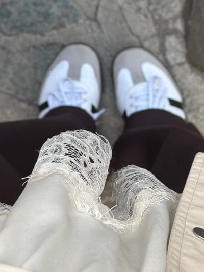 The image presents a downward, close-up perspective of a person standing on a paved surface. The individual is wearing brown leggings, white and grey sneakers with white laces, and a beige jacket. Lace detailing is visible on the edge of the jacket's sleeve. The location appears to be outdoors, evidenced by the stone paving. The scene lacks specific details to confirm the exact location in Lermontovo, Armenia. There is no discernible interaction between the person and any other subject or object.