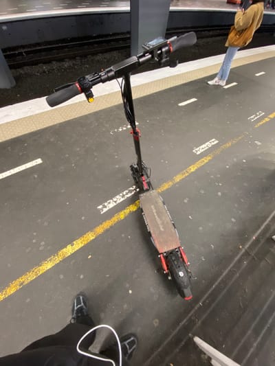 A black and red UrbanGlide E-CROSS PRO electric scooter is centrally positioned on a dark gray asphalt train station platform in Fontenay-sous-Bois, France, likely at either Gare de Fontenay-sous-Bois or Gare du Val de Fontenay. The scooter features a wooden-textured deck, black handlebars with rubber grips, a yellow button on the left grip, and a small digital display. Red accents are visible on its suspension and fender components.

The platform surface displays parallel dashed white lines and a continuous yellow tactile warning strip running along the edge closest to the train tracks, which are visible beyond. A grey concrete support column stands near the tracks.

In the foreground, the feet and lower legs of a person are visible, wearing black pants and black-and-grey sneakers. A white cable rests on the ground near their feet. In the mid-ground, further down the platform, a second individual is partially visible from the waist down, wearing light blue jeans, white socks with a red logo, and dark shoes. They wear a light brown/tan jacket and are holding a dark object in their left hand, facing away from the camera. Both individuals appear to be standing stationary on the platform.