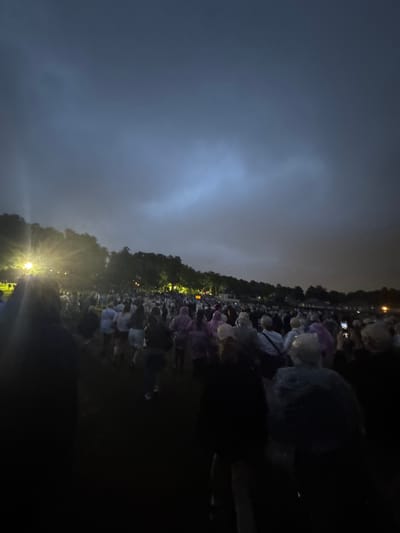 The image shows a large gathering of people outdoors at night, with a dark, cloudy sky. A bright light source is visible on the left side of the image, illuminating the crowd and trees in the background. Many people appear to be wearing ponchos, suggesting the possibility of rain or a concert event. The scene is set against a backdrop of trees and some distant lights, indicating an outdoor public space. The general atmosphere is dark and somewhat dramatic due to the lighting and cloudy sky. The location is described as the city of Glasgow, United Kingdom.