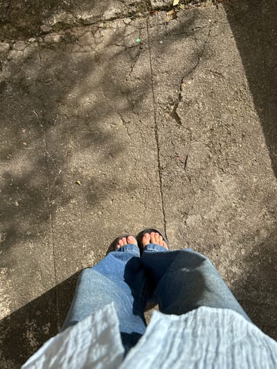 A top-down, full-body shot shows a person wearing blue jeans and dark sandals standing on a cracked concrete surface. Sunlight casts shadows across the ground, highlighting the textures and imperfections of the pavement. The scene appears to be outdoors, possibly in a park or on a sidewalk in Dehiwala, Sri Lanka, given the concrete and natural lighting. There are some small green weeds growing in the cracks of the pavement.