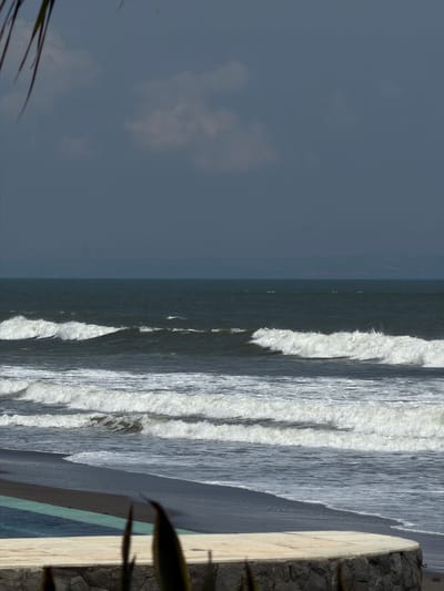 The image captures a coastal scene in Belalang, Indonesia, under an overcast sky. The upper third of the frame shows a dark grey-blue sky with faint cloud formations. Below the horizon, a vast expanse of dark, deep blue ocean displays multiple parallel lines of white-crested waves breaking and rolling towards the shore. The immediate foreground reveals a dark-colored beach where the foamy waves terminate. A curved, low wall constructed of rough, dark grey and brown stones forms the bottom-center and right. Above this wall is a smooth, light-toned curved surface, which abuts a narrow strip of clear turquoise water, indicative of a swimming pool or similar man-made water feature. Out-of-focus dark green, elongated plant leaves are partially visible in the extreme bottom foreground and indistinct dark foliage fringes the top-left corner. No individuals or specific activities are present within the frame.