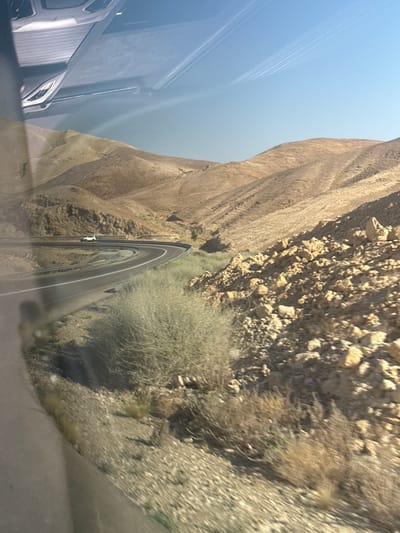 The media file shows a view from inside a vehicle looking out at a winding road carved into a rocky, desert-like hillside. The landscape is characterized by steep, tan-colored cliffs and sparse, dry vegetation. The road appears to be asphalt with white lane markings. A white vehicle is visible driving on the road, navigating a sharp curve. The sky is a clear, pale blue, suggesting it is daytime. Reflections on the window of the vehicle obscure parts of the scene, particularly on the left side where the interior of the vehicle, including a silver car part, is visible. The overall impression is of a scenic drive through a rugged, arid environment. No people are clearly visible, and no text is discernible. The lighting indicates it is likely late morning or early afternoon. The scene is consistent with a location in the Tamar Regional Council, Israel, known for its desert landscapes and winding roads.