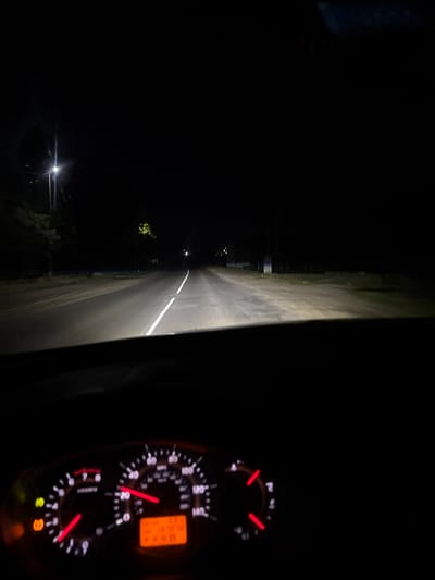 The image depicts a nighttime scene observed from the driver's perspective inside a stationary vehicle in Лабжанскі сельскі Савет, Belarus. The foreground features the illuminated instrument cluster of the vehicle, displaying an odometer/trip meter with an orange digital readout showing numbers and the text "P AND," indicating the transmission is in Park. The speedometer needle points to approximately 30-35 units (likely km/h, given the context of Belarus), while the tachometer indicates engine RPMs are between 1.5 and 2. Several warning lights are illuminated on the left side of the dashboard, including an orange tire pressure warning symbol, a yellow traction control/stability control warning symbol, and a green indicator light (possibly for headlights).
Beyond the vehicle's dashboard, the scene through the windshield shows a two-lane asphalt road extending straight into the darkness, illuminated by the vehicle's headlights and a distant streetlight on the left side. A solid white line marks the center of the road. On the immediate left side of the road, a dirt shoulder is visible, alongside a vertical utility pole with a brightly illuminated light source. Further down the road on the left, faint outlines of trees or vegetation can be discerned. The right side of the road appears to have a grassy shoulder or field, receding into the darkness. The sky is completely dark, indicating a clear night with no visible stars or moon. No people or other vehicles are distinctly visible within the frame.