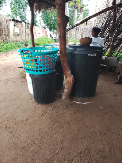 In Barikin Sibre, Nigeria, a scene unfolds outdoors under a thatched roof supported by wooden posts. The ground is covered in sand. In the foreground, a bright teal laundry basket, filled with colorful items, sits atop a black plastic bin. To the right of the laundry basket, a large black water barrel stands, with a brown bowl resting on its lid. A young woman wearing a dark head covering and a white t-shirt is visible behind the water barrel, looking to her right. In the background, a rustic structure with a corrugated metal roof and walls made of mud and woven materials is visible, with dense green foliage and trees surrounding it.