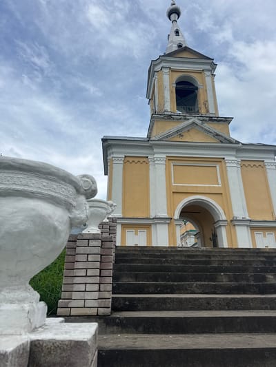 The image shows a low-angle view of a yellow bell tower with white trim, against a cloudy sky. In the foreground, there are concrete stairs leading up to an arched entryway of the bell tower. A large white urn and a brick pillar are visible on the left side of the stairs. The bell tower appears to be part of a religious building, possibly a church or monastery, given its architectural style and the presence of a dome at the very top of the tower. The overall scene suggests an outdoor location, likely in the city of Рассвет, Russia, as indicated. There are no people visible in the image.