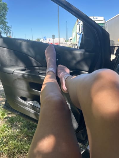 A close-up, low-angle, rotated view shows a person's legs, one bent at the knee, protruding from the open door of a black car. The person is wearing light-colored socks with pink and white stripes. The car door's interior panel is visible. Beyond the car, the background shows a partially visible white truck and street signs under a clear blue sky, possibly located in or near Velburg, Germany, as indicated in the prompt. Grass and vegetation are visible near the bottom of the frame, suggesting the car is parked on the roadside or near a grassy area.