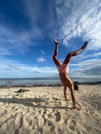 A man is jumping in the air in what appears to be an attempt to do a push-up against the sand on a beach near Sainte-Anne, France. The man is tan, has curly dark hair, and wears pink shorts. His hands are planted on the sand, and his body is suspended horizontally. His legs are extended upward and slightly back. The beach is sandy and extends to the water's edge, where people are standing in the water. In the background is an ocean and the sky contains white clouds against a blue sky.