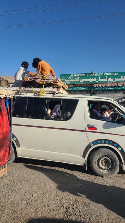 This is an image of a white van parked on a street. Two men are on top of the van, securing wooden planks and other materials.  Two men are in the front seats, and one man is in the back of the van.  A sign for "AL KAREEM PHARMACY KUCHLAK" is visible in the background.  The text on the side of the tire reads "2025". The image was likely taken in Kuchlak, Pakistan.