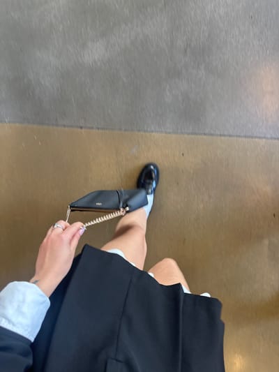 A high-angle view captures the lower body of an individual walking indoors. The person is dressed in a black skirt, white socks, and black patent leather loafer-style shoes on the visible left foot. A white shirt cuff is visible on the left arm, and the left hand, adorned with two rings on the index and middle fingers, holds a small black shoulder bag with a gold chain strap. The floor consists of two distinct horizontal sections: a light brown or tan section in the foreground and a lighter gray, textured section in the background, separated by a straight line. The scene is an indoor environment with no discernible features indicating its specific location within Paradise, United States.