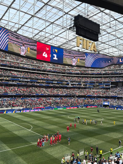 This image captures a wide-angle view of SoFi Stadium in Inglewood, California, United States of America, during a soccer match. The stadium is filled with a large crowd of spectators seated in tiered sections. Above the field, a massive video scoreboard displays a soccer game in progress, showing the score of 4-1 and images of players. The large golden "FIFA" logo is prominently displayed near the scoreboard. The soccer field itself is a vibrant green with white markings. Several players in red and yellow uniforms are visible on the field, engaged in gameplay. A cluster of individuals, likely staff or media, are present near the sideline, some holding cameras. Various sponsor logos, including VISA, Coca-Cola, and Qatar Airways, are visible on banners and around the stadium perimeter. The stadium features a distinctive transparent roof structure.
