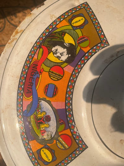 This image depicts a worn, white enameled metal basin or tray, partially seen from a top-down perspective, positioned on a patterned surface, likely flooring, in the city of Isiamigbo, Nigeria. The basin features a decorative, curved decal band with a complex design. The decal includes a black and white stylized portrait of a man with dark hair and a beard, possibly wearing a military cap, set against a yellow background. A red banner displays the word "NIGERIA" vertically in white capital letters. Multiple circular and oval emblems are present, including one prominently labeled "U-STEELS" in yellow text on a blue background. Another oval graphic shows what appears to be a stylized coat of arms with a lion and other symbols. The background of the decal band consists of wavy orange and purple patterns, bordered by a repeating diamond motif in red, green, and blue. The basin exhibits minor rust along its outer edge, particularly at the upper left. Strong natural light illuminates the scene, casting a large, indistinct shadow across the inner right portion of the basin's white surface. No individuals are directly visible, but the depicted portrait represents a person.