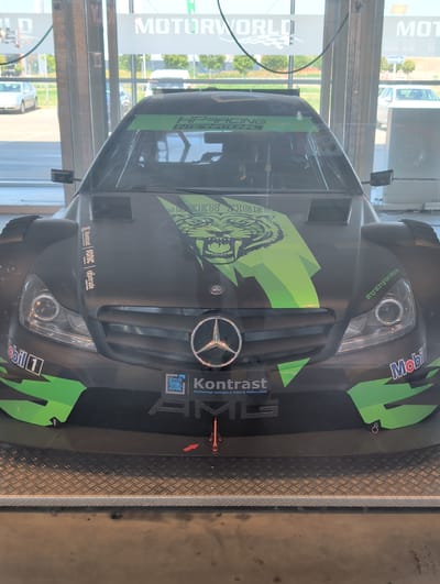 The image shows a black Mercedes-Benz C-Class DTM race car with green accents and various sponsor logos. The car is displayed indoors, possibly in a showroom or museum, with a glass barrier in the foreground. In the background, through the glass, a street scene is visible with parked cars and trees. The text "MOTORWORLD" is visible on a sign in the upper background. The car features the "GREEN TIGER" logo on its hood, along with logos for "Mobil 1," "Kontrast," "HP Racing International," and "#hpracinginternational." The setting appears to be Motorworld Böblingen, Germany, a complex of automotive-themed attractions. There are no people visible in the image. The lighting suggests it is daytime.