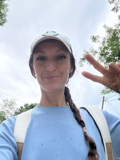 A young woman wearing a light blue crewneck sweatshirt, a light blue baseball cap, and a backpack stands outdoors, looking at the camera. Her right hand is raised, with her index and middle fingers extended in a peace sign. Her hair is dark brown and styled in a braid. The background shows a cloudy sky, green trees, and power lines. There is no information to determine the location in the city of Malyi Mayak, Ukraine.