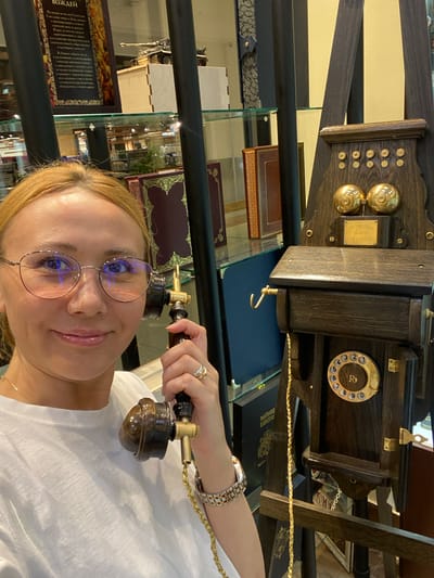 A blonde woman wearing glasses and a white t-shirt is taking a selfie in front of an antique telephone. The telephone is made of dark wood and brass, with two bells at the top and a rotary dial at the bottom. The woman is holding the receiver of the telephone to her ear. In the background, there are shelves with books and what appears to be a model of a tank. The setting seems to be an indoor establishment, possibly a museum or a shop. The overall lighting suggests it is daytime.