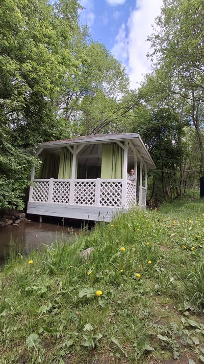 This image, set in Kipen, Russia, captures an idyllic outdoor scene featuring a white wooden gazebo situated beside a narrow body of water, likely a stream or canal. The gazebo, with its decorative lattice railings and light green curtains partially drawn, serves as a focal point.
**Subjects:** Inside the gazebo, a person, appearing to be a man, is visible leaning comfortably on the railing, looking towards the viewer. The gazebo itself is constructed from white painted wood, featuring a gabled roof and intricate cross-patterned latticework on its sides. Lush green foliage from numerous trees surrounds the structure, creating a natural, shaded environment. Above, patches of a bright blue sky with soft white clouds can be seen through the tree canopy. On the right side of the frame, there is an expanse of green grass dotted with vibrant yellow dandelions and other wild plants. The stream flows gently beneath the gazebo and alongside the grassy bank.
**Activity or event taking place:** The man in the gazebo appears to be relaxing and enjoying the tranquil outdoor setting. There isn't a specific event, but rather a moment of leisure.
**Notable details:** The scene suggests a pleasant daytime, likely during spring or early summer, given the fresh green leaves and blooming dandelions. The weather appears clear and sunny. The gazebo is designed for relaxation, offering a shaded spot by the water. No visible text is present in the image. The overall atmosphere conveys peace and natural beauty, characteristic of a countryside or park setting.