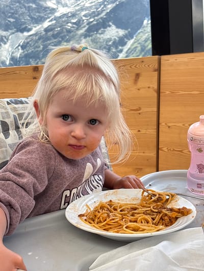 A young child with blonde hair and a purple shirt sits in a high chair, eating spaghetti. The child has sauce smeared around their mouth and is looking at the camera. A plate of spaghetti with a fork is on the table in front of the child. In the background, there is a wooden wall and a window looking out onto a mountain landscape. The setting appears to be a restaurant in Heiligenblut am Großglockner, Austria, given the mountainous scenery. A pink sippy cup is visible to the right of the child.