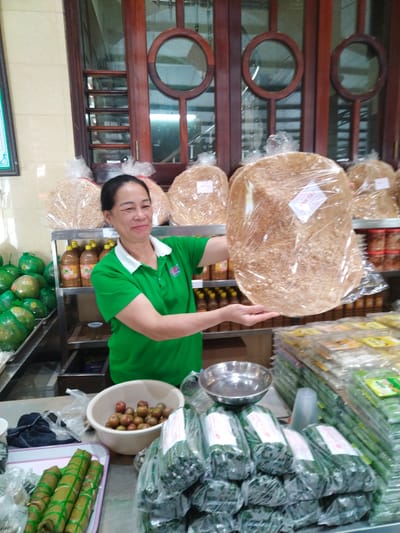 A medium shot captures a middle-aged Asian woman in a green polo shirt with a white collar, standing behind a display counter, in Chu Văn An Ward, Vietnam. She is holding up a large, round, cellophane-wrapped food product, possibly a cracker or cake. Behind her is shelving stocked with bottles of yellow liquid.

On the counter in front of her are several items: a white bowl filled with small, brown, round objects, possibly fruits; stacks of cellophane-wrapped, cylindrical green objects tied with green bands; a metal bowl; and stacks of cellophane-wrapped rectangular objects with green labels. The background features a wooden window with decorative circular glass inserts and additional wrapped circular food products on the shelves behind the woman.