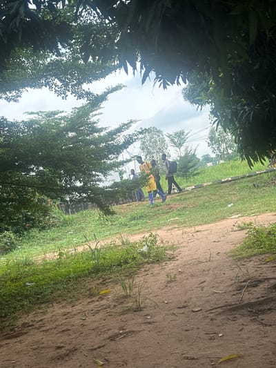 The image depicts a group of four young men walking on a dirt path beside a grassy area in Amansee, Nigeria. The men are casually dressed, with one individual wearing a yellow shirt and blue shorts. They appear to be walking in the same direction. The environment is lush and green, with trees and vegetation surrounding the path. The sky is overcast, indicating a cloudy day. In the background, there are some structures and power lines, suggesting a semi-urban or rural setting. The ground is predominantly dirt, with patches of grass and some scattered debris. The scene appears to be a common day in this Nigerian location.