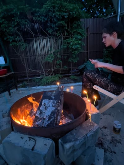 In the dimly lit outdoor setting of Вельяминово, Russia, a young person with dark hair sits on a folding chair, facing away from the camera, towards a bonfire. They are wearing a black top and patterned black lace pants. The bonfire, contained within a metal ring on a bed of sand and surrounded by concrete blocks, burns with bright orange flames and emits smoke. Several lit candles of varying colors are placed on the concrete blocks near the fire. To the right of the scene, a can of Corona Extra is visible on the ground. The background features a dark wooden fence and lush green foliage.