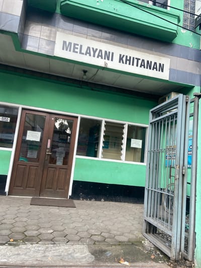The image shows the exterior of a building in Kecamatan Cimahi Selatan, Indonesia (latitude: -6.932, longitude: 107.564). The building is painted a light green color and has a sign above the entrance that reads "MELAYANI KHITANAN." The sign is white with black lettering. There is a double wooden door with a sign that says "BUKA OPEN" located on the left side of the entrance. The entrance is flanked by two windows that are open. The windows have white vertical louvers. A metal gate is on the right side of the entrance. The ground is covered in gray hexagonal pavers. There is a small brown welcome mat located in front of the entrance. The building appears to be a medical clinic or a health facility. There are no people visible in the image. The scene is calm and devoid of any activity. There is no other information available about the scene.