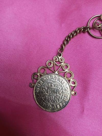 A single, ornate metallic pendant and chain are displayed against a fuchsia pink fabric background. The pendant features a circular, silver-toned medallion, approximately 3 cm in diameter, which is centrally embossed with a crowned emblem and surrounded by script that includes the legible year "1833". This medallion is encased by an intricate, gold-colored filigree frame composed of decorative scrolls and loops. A brass-colored, twisted-link chain extends from the upper part of the pendant, leading upwards and towards the upper right out of the frame. The background consists of a smooth, slightly lustrous fuchsia pink textile, exhibiting minor folds and creases. No individuals, actions, or direct interactions are present within the scene. The location is Qarabucaq qəsəbə, Azerbaijan.