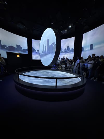 The image shows an indoor exhibit featuring displays of the Chicago skyline. A crowd of people is observing the displays. The displays are circular and rectangular screens showing views of the city. An "EXIT" sign is visible on one of the screens.