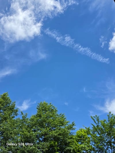 A wide-angle, eye-level shot captures a vibrant blue sky populated with wispy white clouds. The tops of lush green trees fill the bottom portion of the frame, their dense foliage obscuring the ground below. A watermark on the bottom left corner reads "Galaxy S26 Ultra." The scene is outdoors, likely during daytime given the bright sky and clear visibility. The image appears to be taken from West Hartford, United States of America.