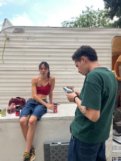 Two individuals are seated on a white corrugated metal surface in front of a white trailer. The person on the left, a woman, is wearing a red halter top and denim shorts. She is looking towards the camera. To her left are three cans and a burgundy bag with a patterned strap. To her right is a can. The person on the right, a man, is wearing a green t-shirt and blue jeans. He is looking down at a piece of folded silver material he is holding in his hands. He has tattoos on his left arm. To his right, a black fan is visible. The scene appears to be outdoors, with trees and a cloudy sky in the background. The trailer has a yellow rope attached to it, hanging down the side. The location is the exterior of a mobile home lot in Sant Cugat del Vallès, Spain.