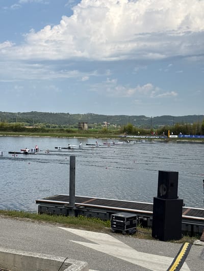 The image depicts a rowing competition on a calm body of water, identified by signage as being at Montemor-o-Velho, Portugal. Several rowing boats with athletes are in motion on the water, navigating designated lanes marked by buoys. In the foreground, a wooden dock structure with metal poles and attached equipment, including large speakers and a road case, is visible. The background reveals a tree-lined landscape with a distinctive square tower structure and a prominent banner reading "MONTEMOR-O-VELHO" and "finish". The sky is partly cloudy.