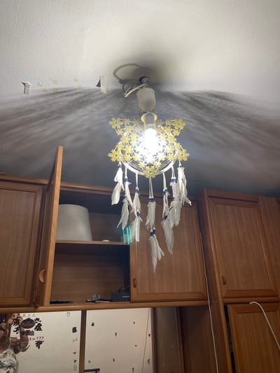 The image shows a ceiling light fixture with a dreamcatcher attached, hanging above a set of wooden cabinets. The light casts intricate shadows on the white ceiling. The cabinets appear to be part of a kitchen or storage area. The dreamcatcher has white feathers and blue beads. The scene is indoors, likely in a residential setting in Santa Coloma de Gramenet, Spain, though no specific landmarks or indicators of this location are visible. There are no people present in the image.
