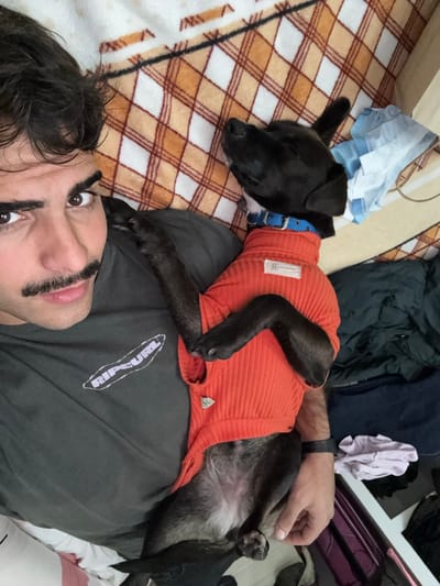 A man is lying on a bed with a dog on top of him. The man is looking at the camera. The dog is wearing an orange sweater and a blue collar. The bed has a brown and white checkered blanket. The location is Sarandí, Argentina. There are clothes and a bag on the bed. The man is wearing a gray shirt. He has dark hair and brown eyes. He has thick eyebrows and a mustache drawn on his face.