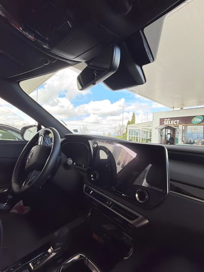The image, taken from inside a car, shows the view through the windshield toward a Shell Select and Shell Cafe gas station. The sky is partly cloudy. The car's interior is primarily black, with the steering wheel and dashboard prominently visible. The location appears to be in the city of Dragor, Bulgaria, based on the signage of the Shell gas station. There are no people visible inside the car. A person can be seen entering the cafe.