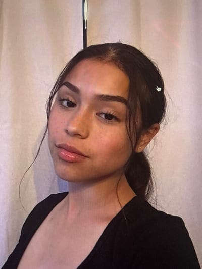 A young woman with brown hair, a few freckles, and glossy lips looks directly at the camera. She wears a dark shirt, and a small silver hair clip is visible in her hair. Her expression is slightly pensive. The background appears to be a white surface, possibly fabric or a wall. The image has a soft, slightly blurry quality.
