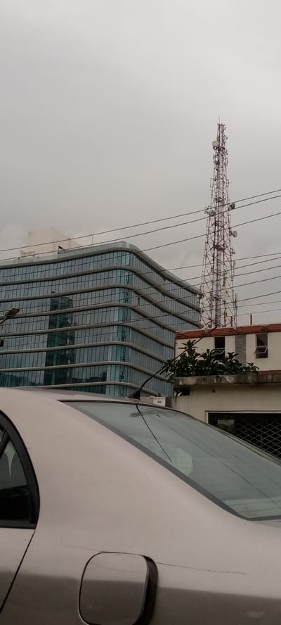 The image captures a scene in Ikoyi, Nigeria, dominated by a large, modern glass-fronted building with a curved facade. Adjacent to this building is a lower, cream-colored structure with open windows and an antenna tower rising behind it. The sky is overcast and gray. In the foreground, the rear section of a light-colored car is visible, with its rear windshield and part of the bodywork in focus. Power lines crisscross the sky above the buildings. A tall telecommunications tower, with multiple antenna dishes, stands prominently in the background, to the right of the modern building. Vegetation is present on the roof of the cream-colored building, and a slender, dark antenna or rod extends from the car's rear windshield towards the left. The overall impression is of an urban environment under a cloudy sky.