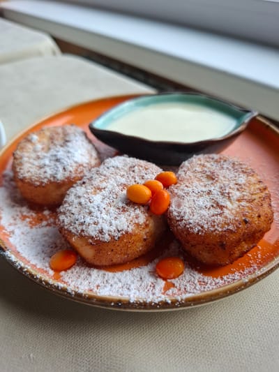 The image shows a plate of syrniki, a type of fried curd cheese pancake, dusted with powdered sugar and garnished with sea buckthorn berries. A small dish of white sauce, likely sour cream or a sweetened condensed milk, is placed behind the syrniki. The plate is orange and sits on a beige tablecloth. The background is blurred, showing what appears to be a windowsill with a view of a building. The scene is lit from the side, casting soft shadows. There are no people or actions depicted in the image, and the location in Sukhoy Ruchey, Russia, is inferred from the context and not visually evident.
