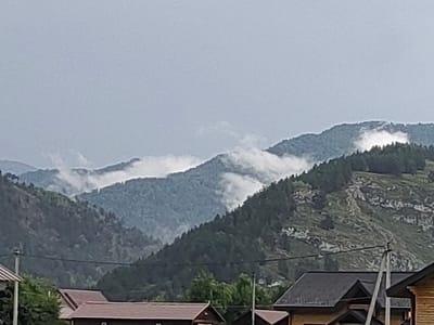 The image depicts a mountainous landscape with a cloudy sky. The foreground shows the roofs of several buildings, possibly houses, with a power line pole visible between them. The mountains are covered in dense green vegetation, and some areas are shrouded in mist or clouds. The overall scene suggests a rural or mountainous area, likely in Anos, Russia, given the context. No people or specific actions are discernible in the image.