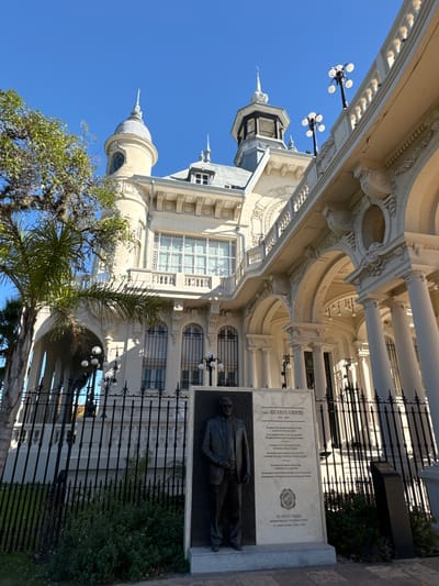 The image is a street-level view of the Museo de Arte Tigre (Tigre Art Museum) in Tigre, Argentina, taken in daylight under a clear blue sky. The museum's architecture is predominantly white, featuring intricate detailing, columns, arches, and a distinctive dome. A monument with a dark-colored statue and inscription is positioned in front of the building, bordered by a black wrought-iron fence and greenery. Street lamps line the area, and some trees are visible, partially obscuring the building. The perspective of the photo is slightly tilted.