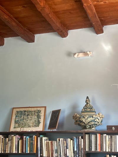 The image shows a room with a light blue wall and a wooden ceiling with exposed beams. On the wall, there is a light fixture. Below the light fixture, on a shelf, there is a framed abstract painting, a smaller dark framed object on an easel, and a decorative urn. The shelf is part of a wooden bookshelf filled with books. The room appears to be a study or living area, possibly in a historical building given the exposed beams. The scene is static and depicts an interior domestic setting. There is no information in the image that directly links it to Militello in Val di Catania, Italy.