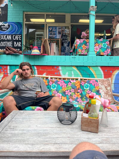 A man sits at a wooden table with a bottle in his hand. The table also contains a basket, a clear container, and a black metal object. He sits in front of a building with a sign that reads "MEXICAN CAFE SMOOTHIE BAR." The wall behind him is painted with a vibrant floral pattern. There is another floral pattern on the cushions of the chairs at the table. The building is painted turquoise.