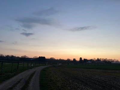 The image captures a semi-rural landscape scene in Würselen, Germany, at either dusk or dawn. The sky, occupying the upper two-thirds of the frame, exhibits a gradient transitioning from deep blue at the top, through lighter blue and pale yellow, to an orange-pink hue at the horizon. Sparse, dark grey clouds are visible in the upper portion, and a slender white crescent moon is present in the upper center-right. A faint, horizontal linear streak is discernible in the mid-right sky near the horizon.
The lower third of the image features a winding, unpaved dirt path that curves from the lower-left foreground towards the center-right background. To the left of the path, a green, grassy paddock is enclosed by a wooden fence consisting of vertical posts and horizontal rails. To the right of the path, a section of dark, tilled earth with visible furrows extends into the midground. A dense treeline, composed of largely bare deciduous trees, forms the horizon. Several structures are visible amidst or behind the trees, including a light-colored house with a dark roof and multiple barn-like buildings with lighter roofs, consistent with agricultural or equestrian premises. No individuals are present in the scene.
