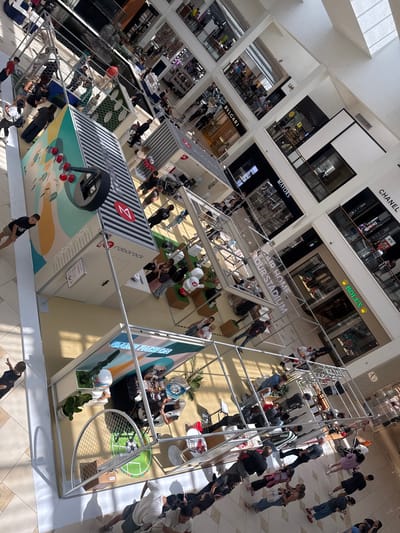 The image depicts a bustling interior scene of the Aventura Mall in Aventura, Florida, United States of America. The perspective is from above, looking down at a multi-level shopping mall with a central atrium. The architecture features a modern design with glass railings and metallic structural elements.
Several temporary exhibits or booths are set up on the ground floor. One prominent display features a large graphic of robots and a stylized wheel, with the text "roborock" visible. Another exhibit has the text "GAME FACE ON" and displays what appear to be gaming-related items or a setup. People are seen milling around these exhibits, some interacting with displays, others observing.
The mall is populated with a diverse group of individuals, including adults and children. They are dressed in casual summer attire, suggesting warm weather. Many are walking, standing, or engaged in conversations. The lighting suggests it is daytime, with sunlight filtering through the mall's upper levels.
In the background, various high-end retail stores are visible, with their prominent signage such as "BVLGARI," "MAYO'S," and "CHANEL." The overall atmosphere is one of commercial activity and social gathering.