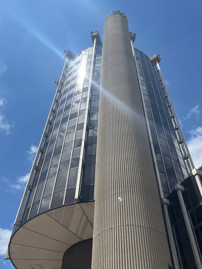 The image shows a low-angle, upward view of the Crédit Lyonnais building in Créteil, France. The building is a modern high-rise with a glass facade and a cylindrical concrete structure running vertically up its side. The sky is a clear blue with scattered white clouds. Sunlight glints off the building's windows, creating lens flare. The architectural style is characteristic of the late 20th century. There are no people visible in the image. The overall impression is one of urban architecture and a bright, sunny day.