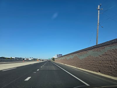 This is a first-person, forward-facing view from a vehicle traveling on a multi-lane highway in West Sacramento, California, United States of America. The highway has asphalt paving with white lane markings and a concrete barrier on the left. On the right, there is a textured brown retaining wall with a diagonal band of grey blocks. A wooden utility pole with wires stands against a clear blue sky. Distant buildings, some with advertisements on them, are visible on the left side of the highway. There are a few cars on the highway, all moving in the same direction as the viewer. The scene appears to be during daylight.