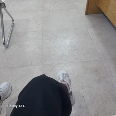 The image presents a high-angle view looking down at a person's lower body and feet in an indoor setting. The main subjects are two feet clad in white sneakers and dark (likely black) pants. The left foot's sneaker shows a glimpse of a pink sock or inner lining, while the right foot's sneaker has a visible textured brown sole.

The floor is made of light-colored, possibly beige or light grey, marbled tiles with visible grout lines. In the upper left corner, a light-colored wooden surface, resembling a table or piece of furniture, is visible. In the bottom left, a shiny, metallic object with a triangular shape, potentially the base or leg of a chair or stand, rests on the floor.

The activity depicted is simply the person standing or moving on the tiled floor. The setting appears to be a basic indoor environment in Nuseirat, Palestinian Territory. No specific time of day or weather conditions are discernible from the indoor lighting. There are no overt emotional cues. The text "Galaxy A14" is visible as a watermark running vertically along the right side of the image, indicating the camera used.