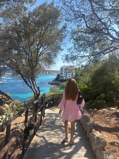 The image depicts an outdoor, sunlit coastal scene in Cas Català-ses Illetes, Spain, overlooking a bay. A single individual, viewed from behind, with long reddish-brown hair, walks downhill on a paved pathway. The person wears a light pink, short, ruffled dress or tunic and dark flip-flops, carrying a dark shoulder bag on the left.
To the left of the path, a wooden railing with attached outdoor lights runs parallel to the descent, beyond which rocky terrain with agave plants and other scrub vegetation slopes towards the water. A large olive or similar tree with light green foliage stands prominently on this side. To the right of the path, the ground is natural, rocky, and covered with low-lying green bushes and trees. Overhead, pine trees frame the bright, clear blue sky.
The pathway leads towards a calm bay with distinct turquoise and deeper blue waters. Numerous boats, including sailboats and motorboats, are anchored in the deeper sections of the bay. Orange buoys or markers are visible in the shallower, turquoise areas. On the opposite side of the bay, a multi-story white building with balconies is situated on a cliff overlooking the water, amidst dense green vegetation. Distant landmasses and subtle structures are visible across the bay. The scene is illuminated by strong natural sunlight, casting clear shadows on the pathway.